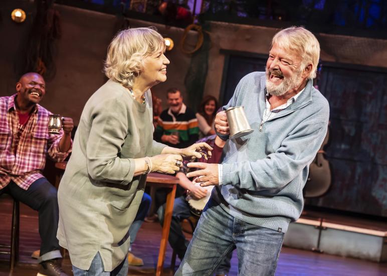 A man in a blue jumper and a woman wearing green laugh together, the man holds a tankard. 