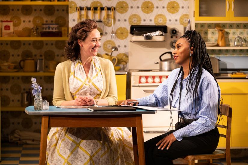 Two women sit talking at a kitchen table, one woman is in a blue pinstripe shirt and the other is wearing a yellow dress and cardigan