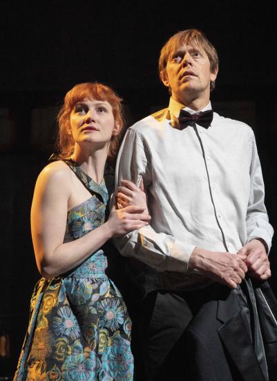 A man and a woman stand arm in arm, he is wearing a tuxedo and she is wearing a green evening gown with flowers on