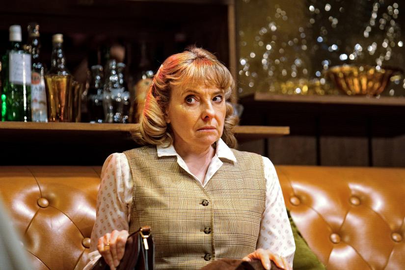 Susan, a woman who looks very uncomfortable, sits on a sofa in a shirt and waistcoat