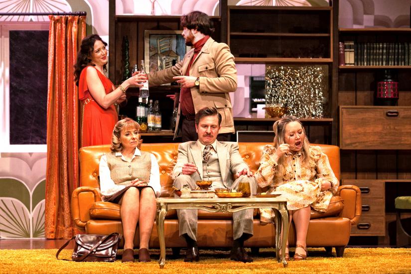 Beverly and Tony flirt, standing behind the sofa. Laurence, Angela and Susan sit in on the sofa looking unhappy