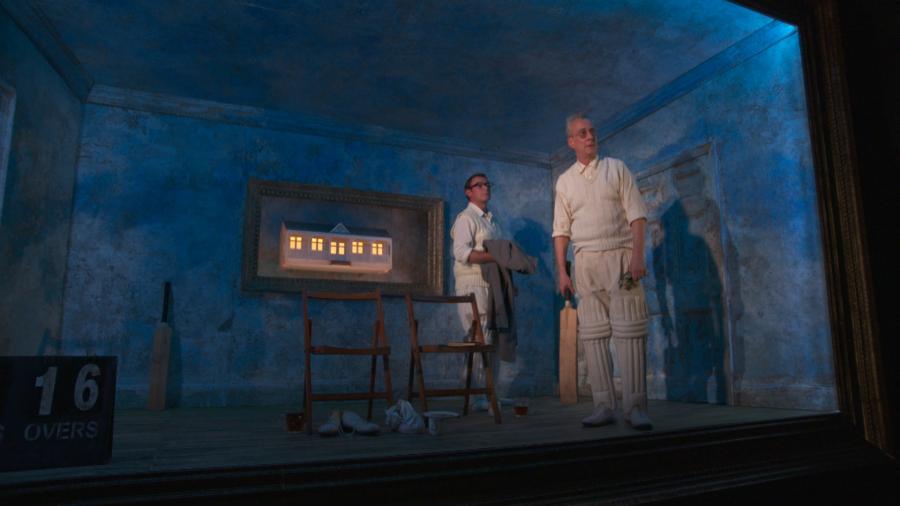 Two men are standing in a blue room, looking out towards the audience. On the back wall there is a small house lit up.