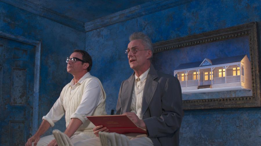 Two men sit together, one holds an open book, they are both wearing cricket gear