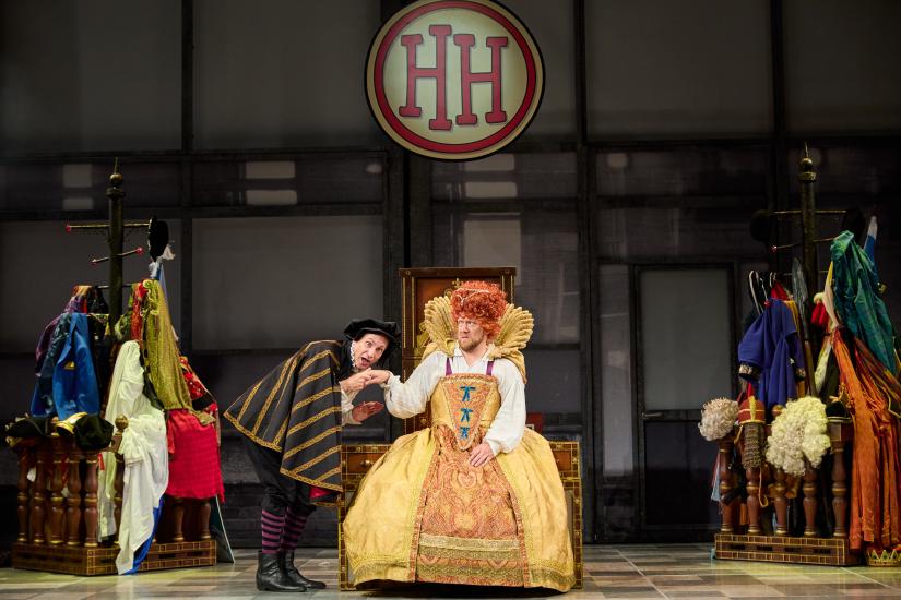 A man bends to kiss the hand of someone dressed as Queen Elizabeth, sitting in a throne