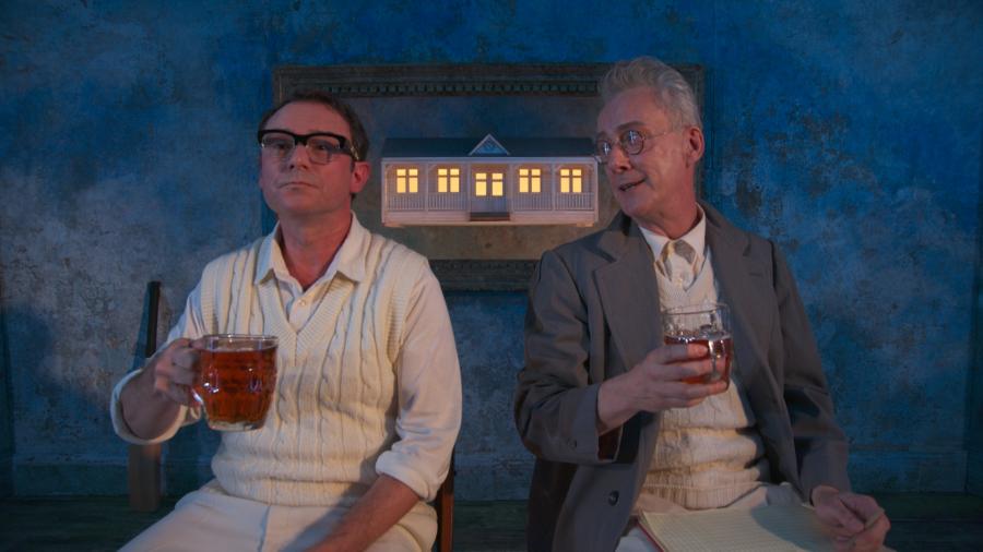 Two men sit together, they are both drinking pints of beer, there is a small lit up house in the background behind them