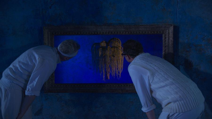 Two men are hunched over looking through a gap in a blue wall at a mysterious object
