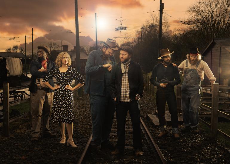The cast of Of Mice and Men stand against a sunset with a railroad track beneath their feet