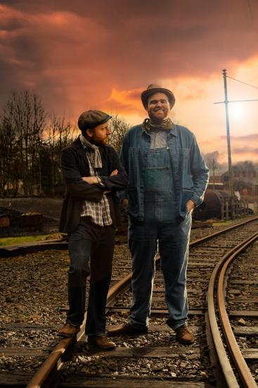 George and Lennie stand together on a railroad track, sun sets behind them