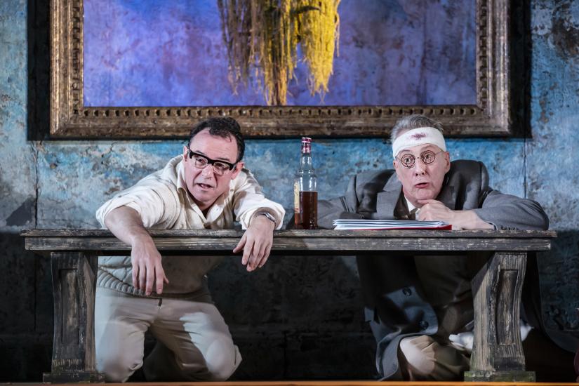 Two men lean against a bench with an open bottle of whisky between them. One is wearing cricket whites and the other has a bloody bandage wrapped around his forehead.