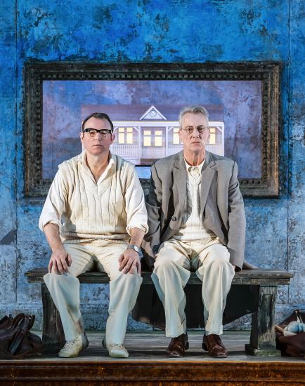 Two men wearing cricket whites sit together on a bench, there is a lit up house behind them