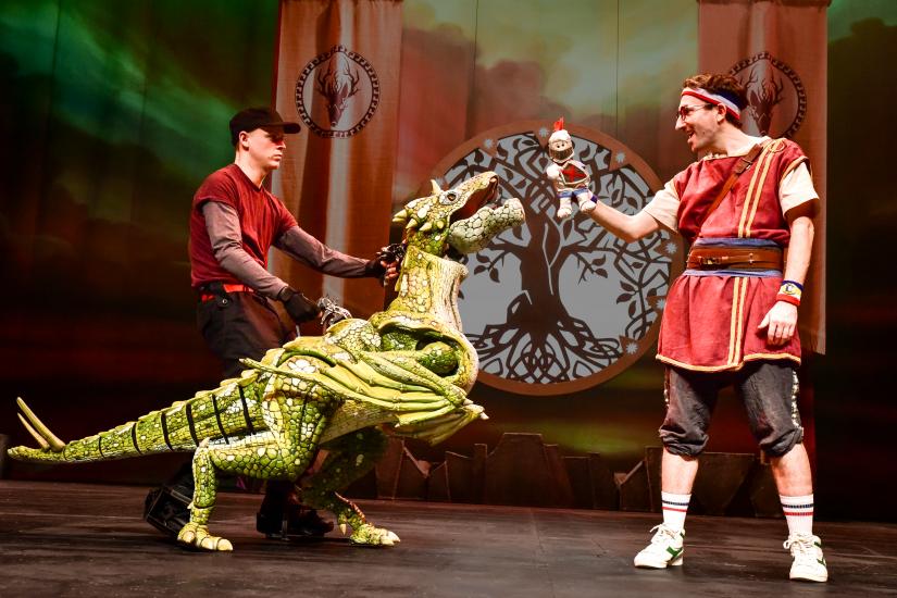 A man stands in front of a green dragon, holding a small puppet in his right hand