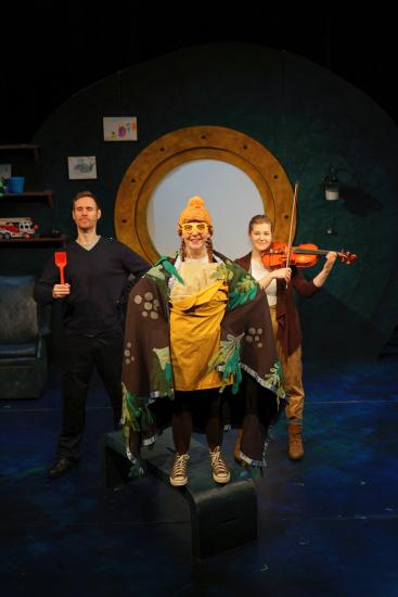 A girl stands with a yellow beanie, yellow goggles and cape on, behind her are a woman playing the violin and a man holding an orange spade