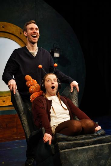 A girl sits in an armchair with an orange snail shell and antenna on her head, a man stands behind the chair smiling 