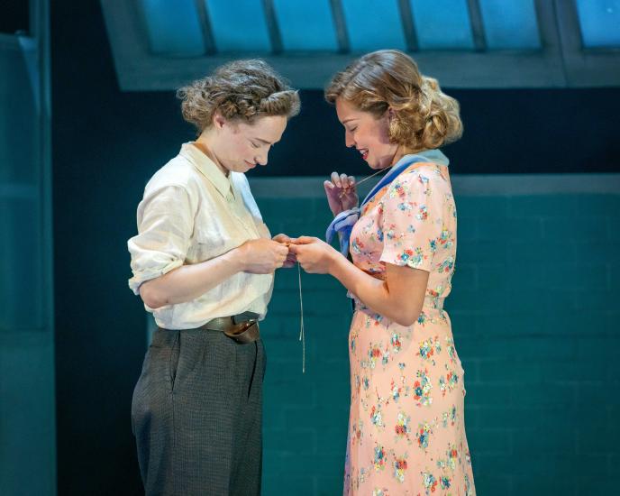 Two women stand together looking at a necklace, one has a floral dress on and the second is dressed in a shirt and trousers