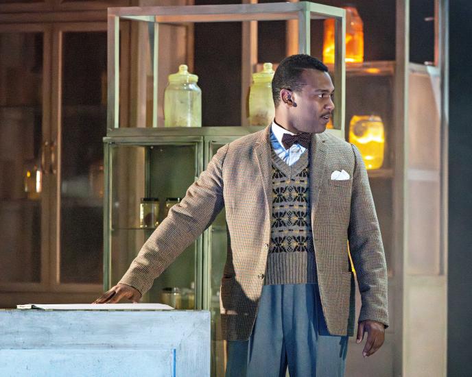 A man stands wearing a brown tweed jacket, sweater vest, bow tie and blue