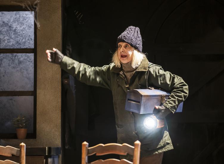 A woman in a wax jacket and a knitted beanie hat is carrying a torch and a toolbox, she has her arm outstretched and looks shocked