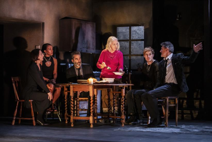 Five people dressed in dark suits and clothing sit around a dining table by candelight, whilst a woman in a red jumper serves them food