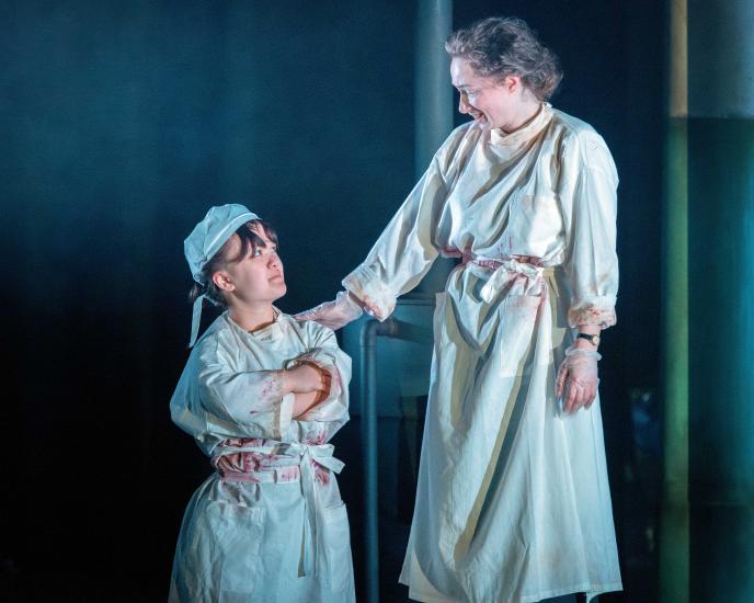 Two women stand together wearing bloodied doctors coats, one has her arms crossed and the second has a reassuring hand on her shoulder