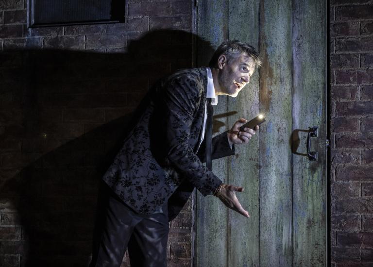 A man in a dark suit stands against a brick wall and wooden door, he is holding up a smartphone to use as a torch