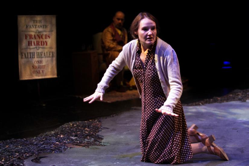 A woman kneals on the floor, she has her hands by her sides with her palms flat. She wears a brown dress.