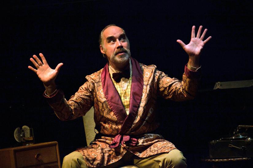 A man with a moustache sits on a chair, his arms are up and his hands are raised with palms facing the camera