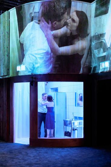 Mike Noble as Macbeth, and Laura Elsworthy as Lady Macbeth. They stand in a room to the side of the carpeted stage, and we see them talking intensely through a large glass window. We see the CCTV footage of this in close up on a screen above them.