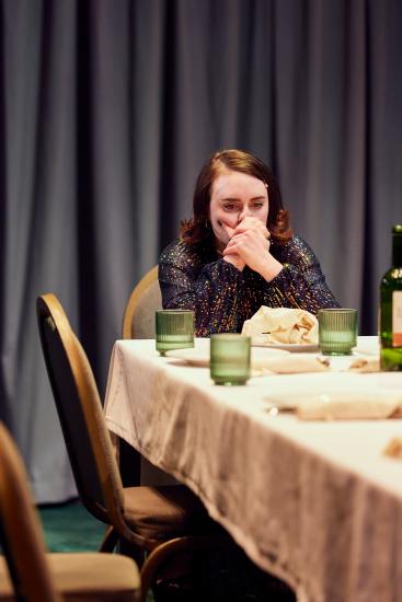 Laura Elsworthy as Lady Macbeth, sits at the head of a banquet table in a glamorous sequin dress with her hands to her mouth looking concerned. 