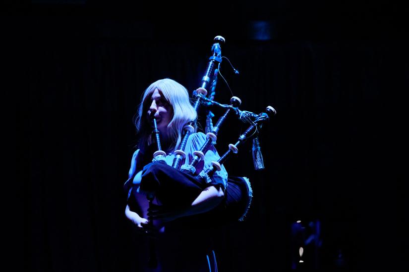 Coral Sinclair as an apparition,  wearing a blonde wig and blue dress playing the bagpipes in dramatic lighting. 