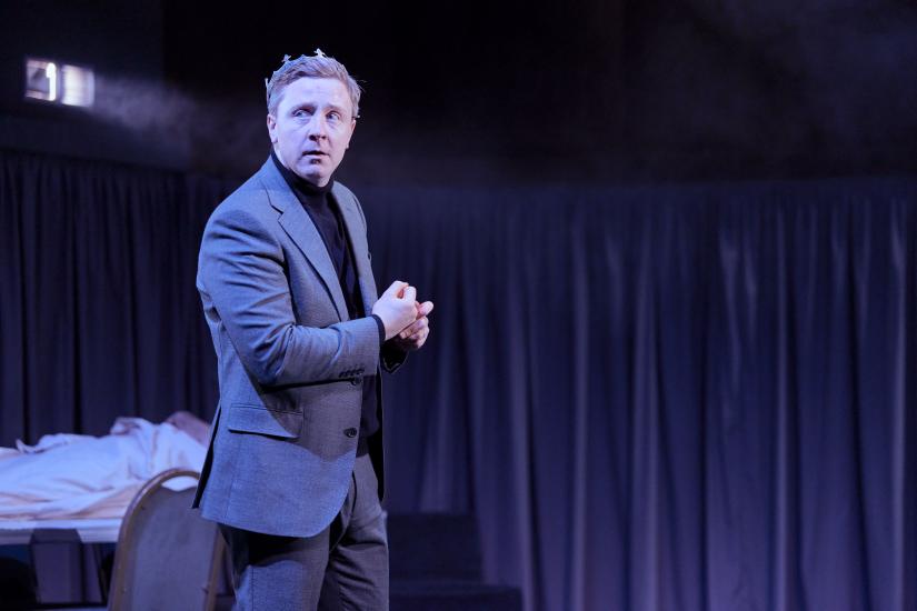Mike Noble as Macbeth wears a smart grey suit and a crown on his head. 
