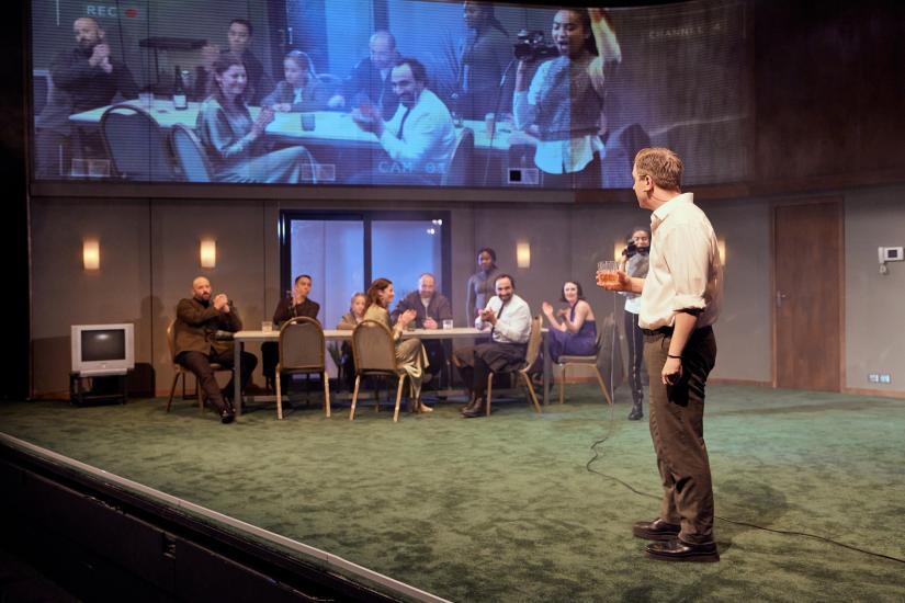 The cast of Macbeth sit around a banquet table as Malcolm, played by Jasmine Elcock films them on a Go-pro camera, which is being played on the screens above the stage. 