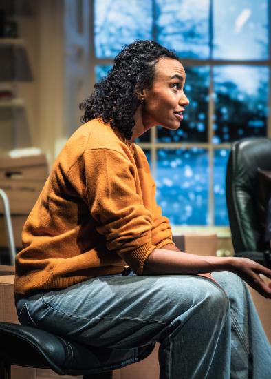  A woman sits in a chair, she is wearing jeans and an orange jumper