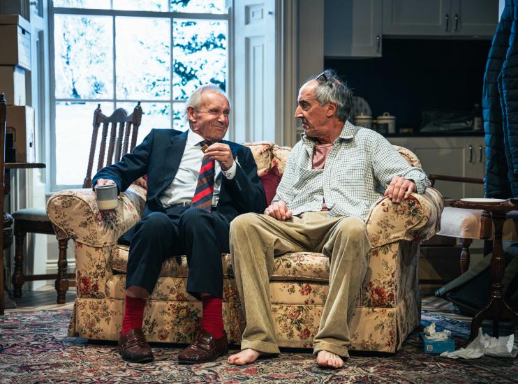 Two older man sit on a sofa, one has a cup of tea and a biscuit, the other looks dishevelled