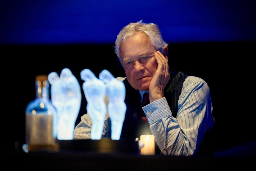 A man in a shirt and waistcoat sits with his head in his hand looking at four glass figurines of a human body