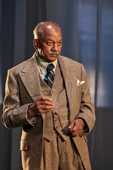 A man wearing a brown tweed suit and waistcoat is holding a glass tumbler