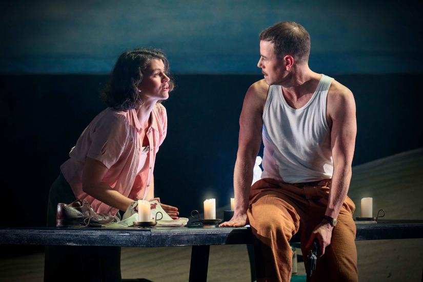 A woman and a man talk, stood either side of a large desk with candles, papers and a brown shoe on it