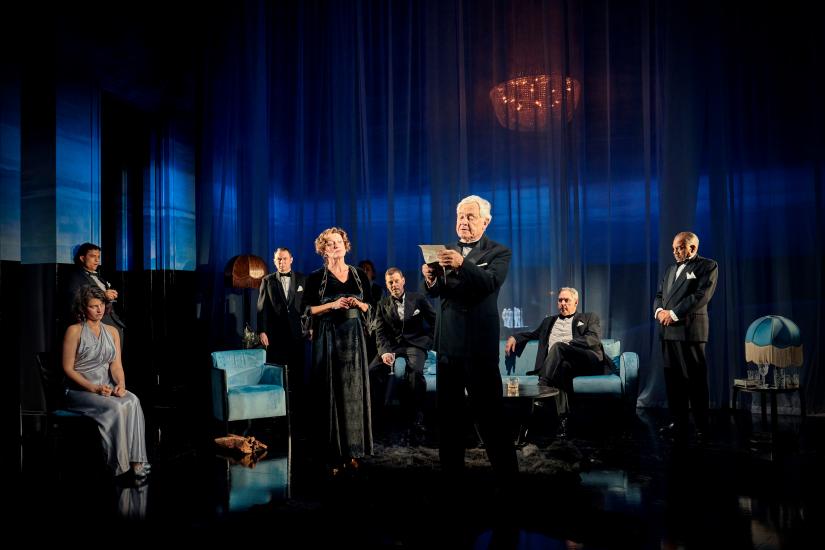 A group of people stand and sit in a grand living room, all wearing evening clothes. A man stood in the centre is reading a letter