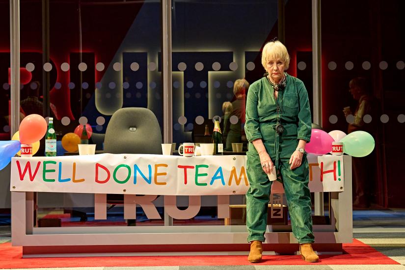 A woman leans against a table with a party banner across it, she looks glum