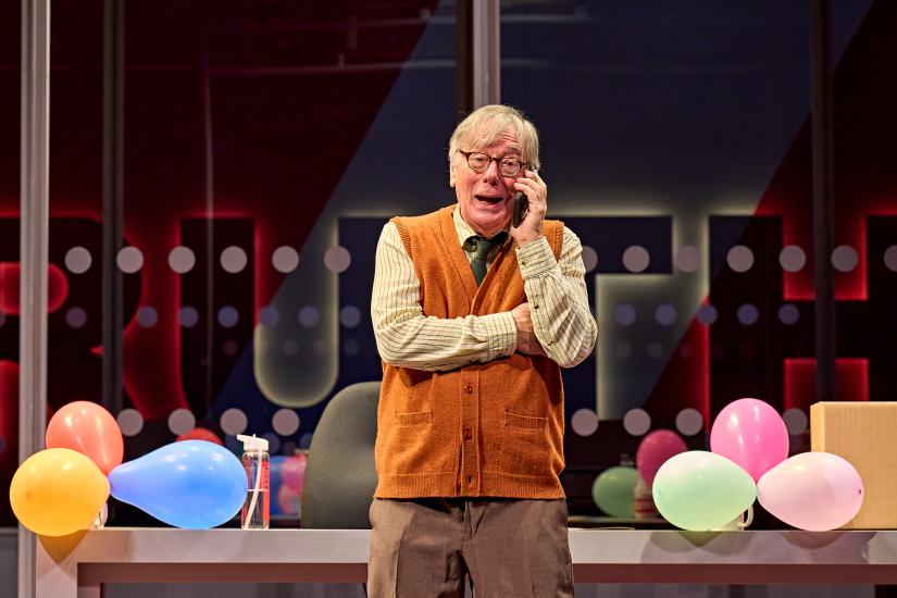 A man in a brown vest and shirt holds a phone to his ear