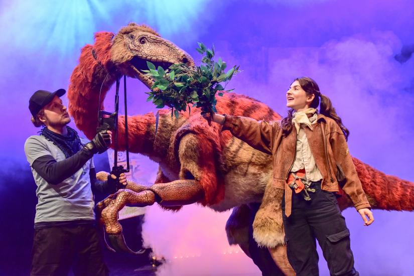 A dinosaur eats some leaves from a branch held by a woman