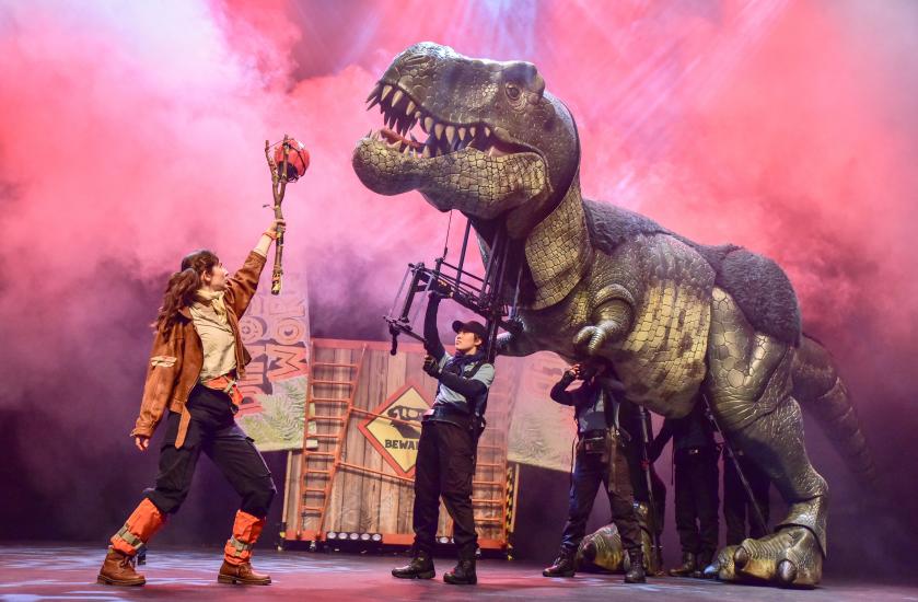 A t-rex tries to get meat from a stick held by a woman