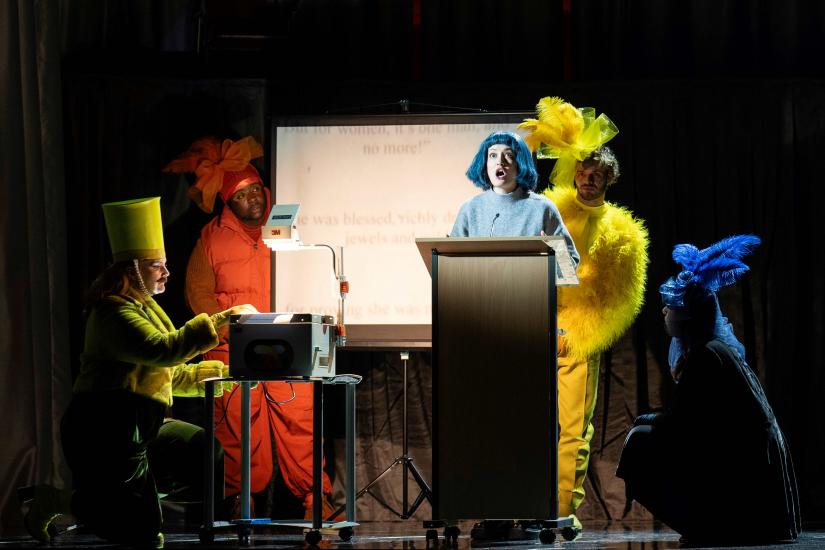 Blue haired woman stands at podium in classroom while other characters dressed in different colours look on