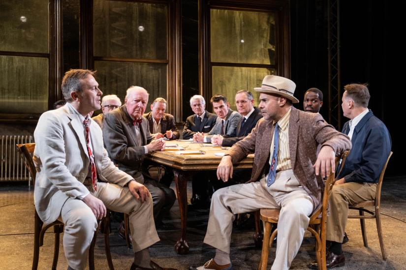 Twelve male jurors sit around a table in discussion