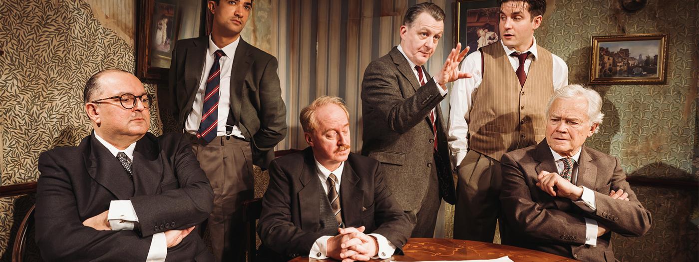 Six men in suits stand and sit by a table in discussion