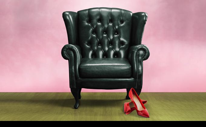 A black chesterfield armchair with a pair of red high heels on the floor in front of it