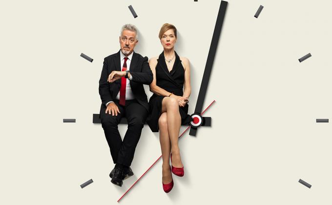 A man and woman sit on a clock face, they both look fed up