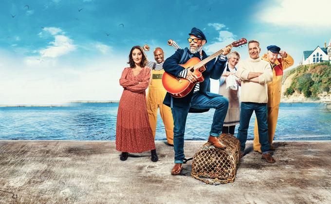 A fisherman with a guitar stands in front of the sea with a group of people
