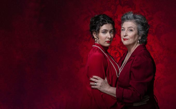 A woman and her daughter embrace, both wearing red dresses and pearl necklaces