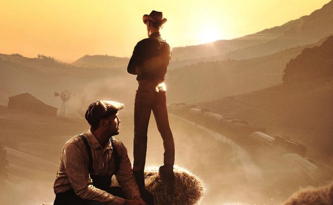 One man stands, another sits, against a backdrop of an American dusty landscape. One man wears a cowboy hat.