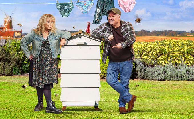 A woman and a man lean against a beehive, in the background there are fields and a washing line