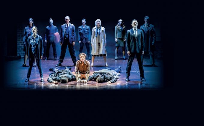 A group stand in the middle of a stage, two men lie on the ground, one each side of a woman who looks sorrowful